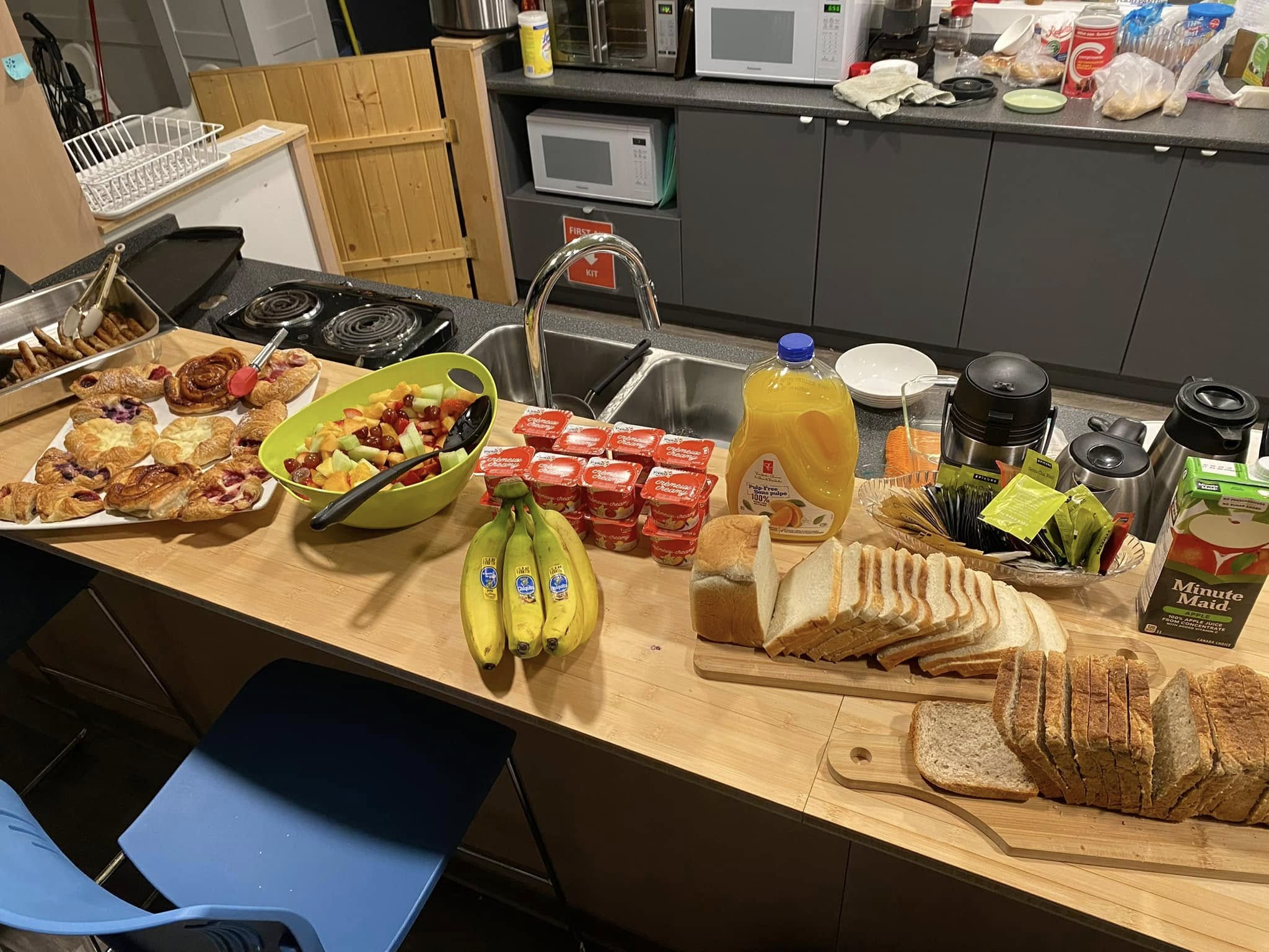 A table set with breakfast foods including sliced bread, pastries, bananas, yogurt cups, fruit salad, juice, and hot drink carafes arranged in a kitchen space with a sink and appliances in the background.
