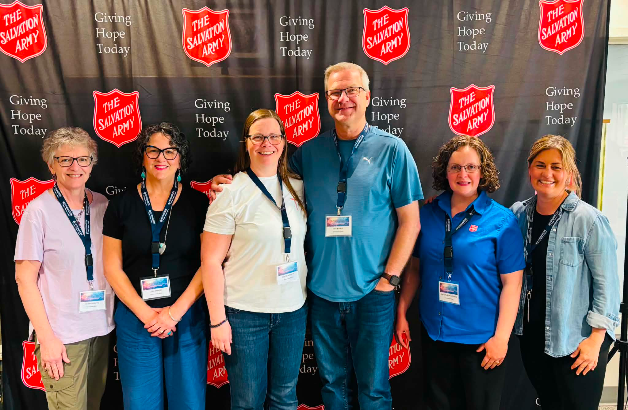 Seven people wearing conference name badges stand together in front of a black backdrop with repeated Salvation Army shield logos and the words Giving Hope Today, posing for a group photo at a Compass Conference event.