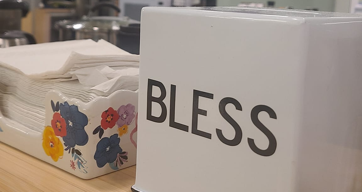 A stack of folded white napkins in a decorative holder with a floral pattern sits beside a white container labeled BLESS in bold letters on a wooden countertop.