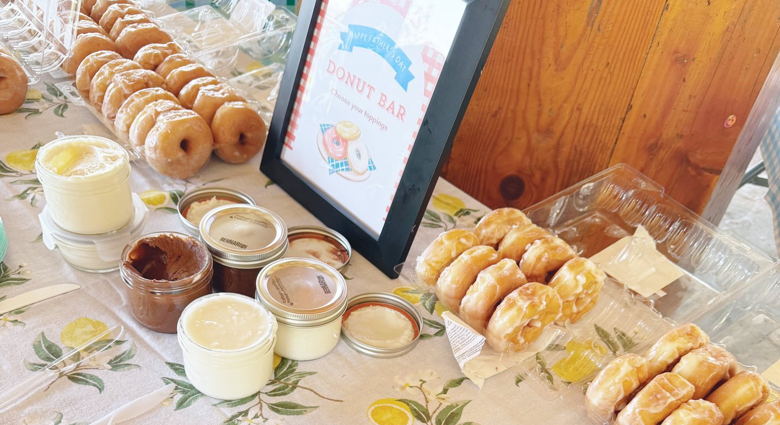 Table display for a Fathers Day donut bar featuring rows of glazed donuts, small jars of toppings and spreads, plastic utensils, and a framed sign reading Happy Fathers Day Donut Bar, Choose your toppings.