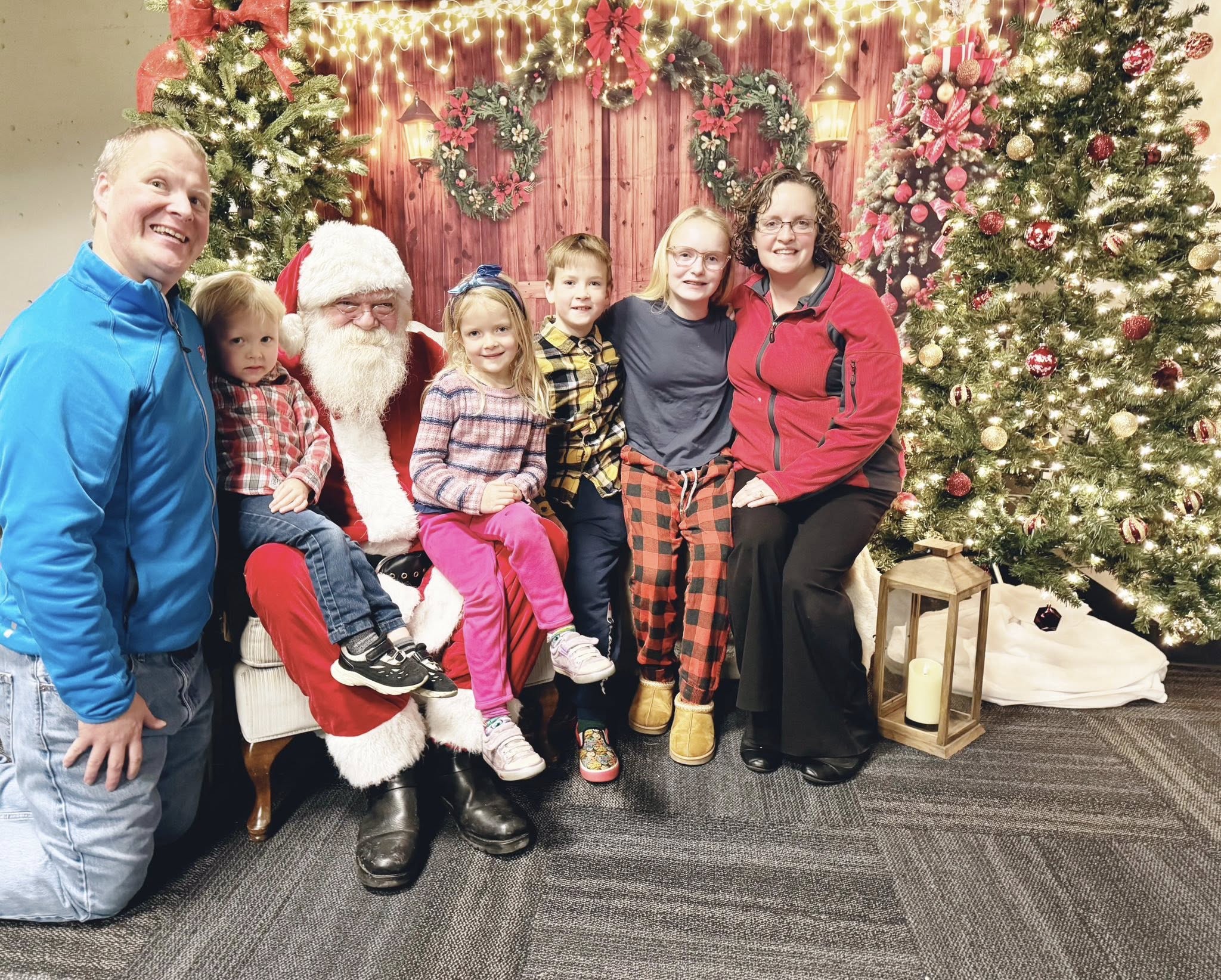 Group of adults and children seated and standing together with a person dressed as Santa Claus in a festive indoor setting, surrounded by decorated Christmas trees, wreaths, and holiday lights.