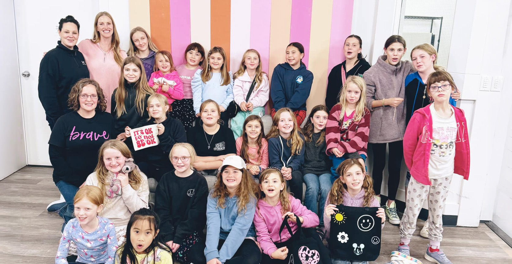 Group of children seated and standing together with two adult leaders in front of a colorful striped wall, posing for a group photo during a Brave Junior program activity.