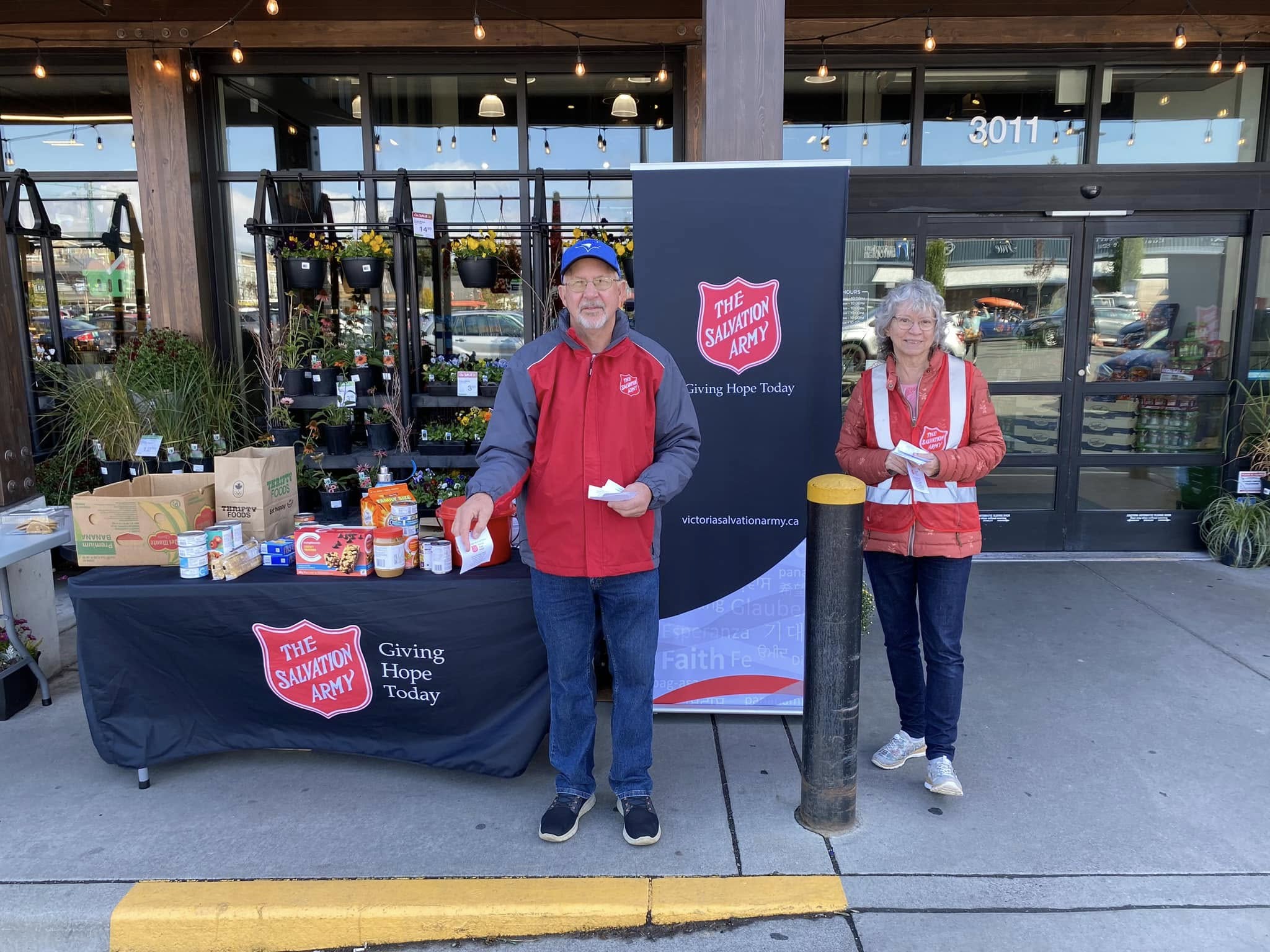 Two Salvation Army volunteers stand outside a storefront beside a display table with donated food items and a banner reading ‘The Salvation Army – Giving Hope Today,’ with plants and store entrance doors visible in the background.