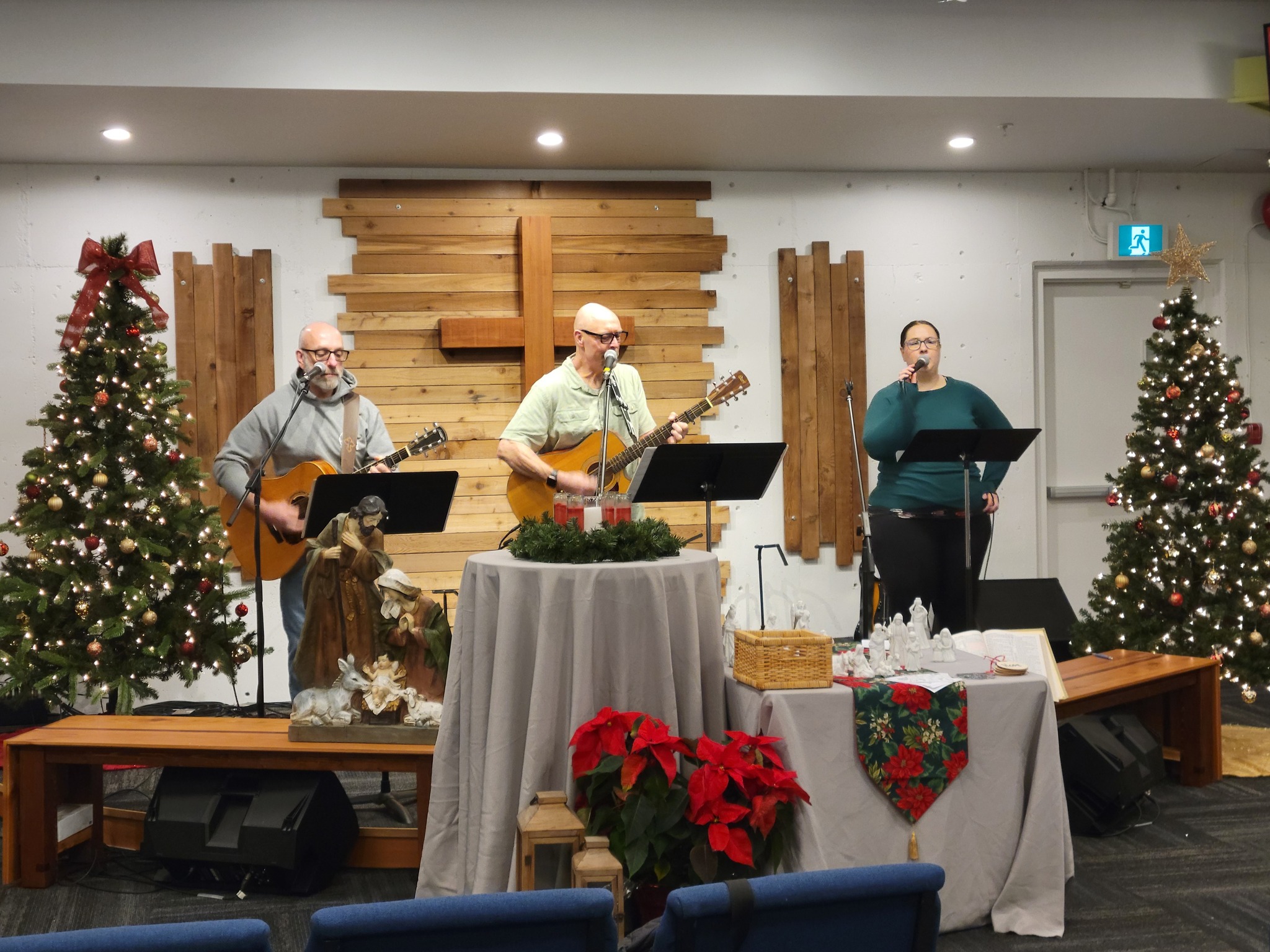 Worship team leading a church service with guitars and microphones at the front of a sanctuary, with a wooden cross backdrop and Christmas decorations including trees and seasonal displays.