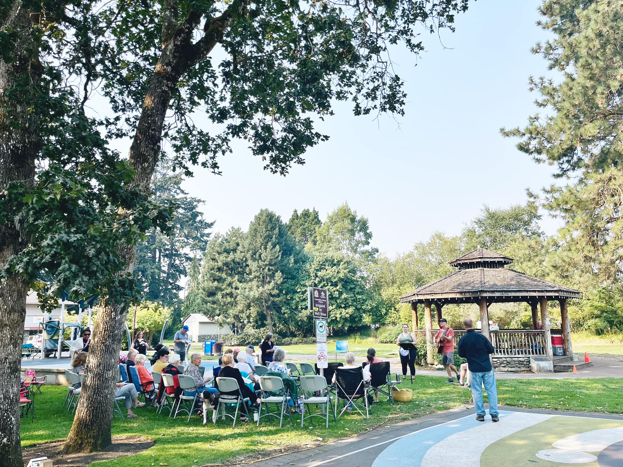 People gathered for an outdoor worship service in a park, seated on folding chairs facing a speaker area, with trees, open green space, and a gazebo nearby.