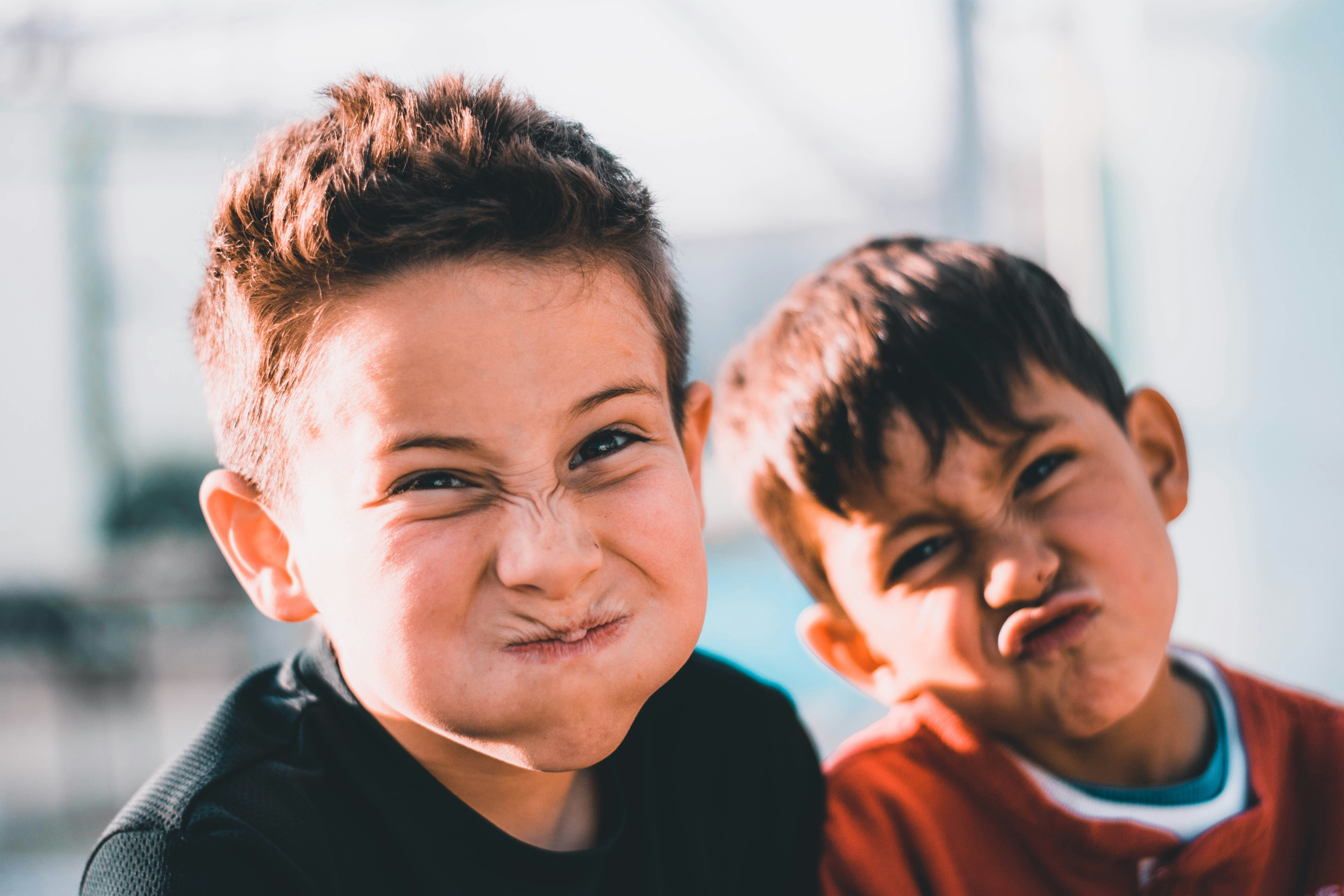 Two children close to the camera outdoors, making exaggerated facial expressions while standing side by side in natural light.