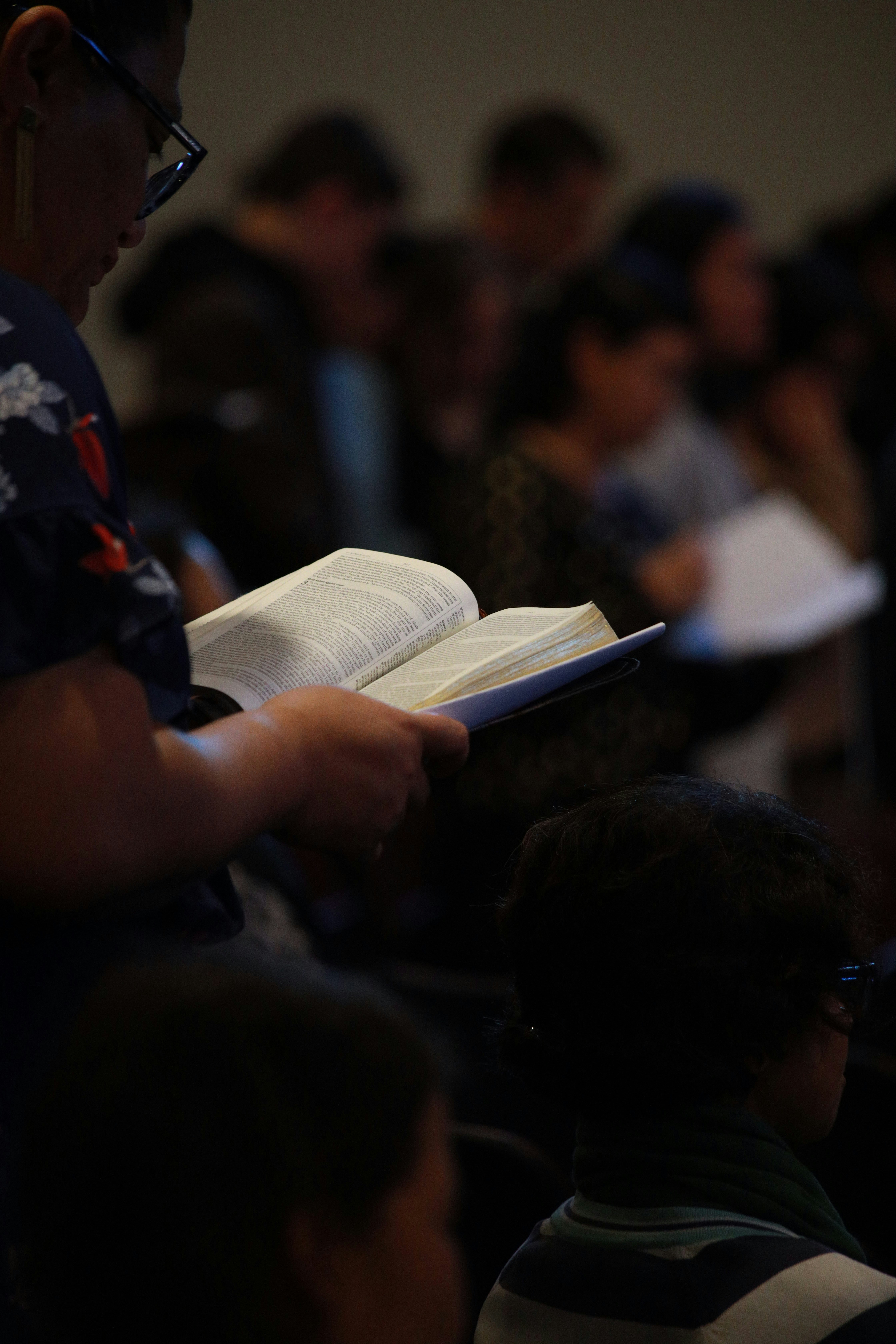 Close-up of hands holding an open Bible in a dimly lit indoor setting, with other seated people softly out of focus in the background.