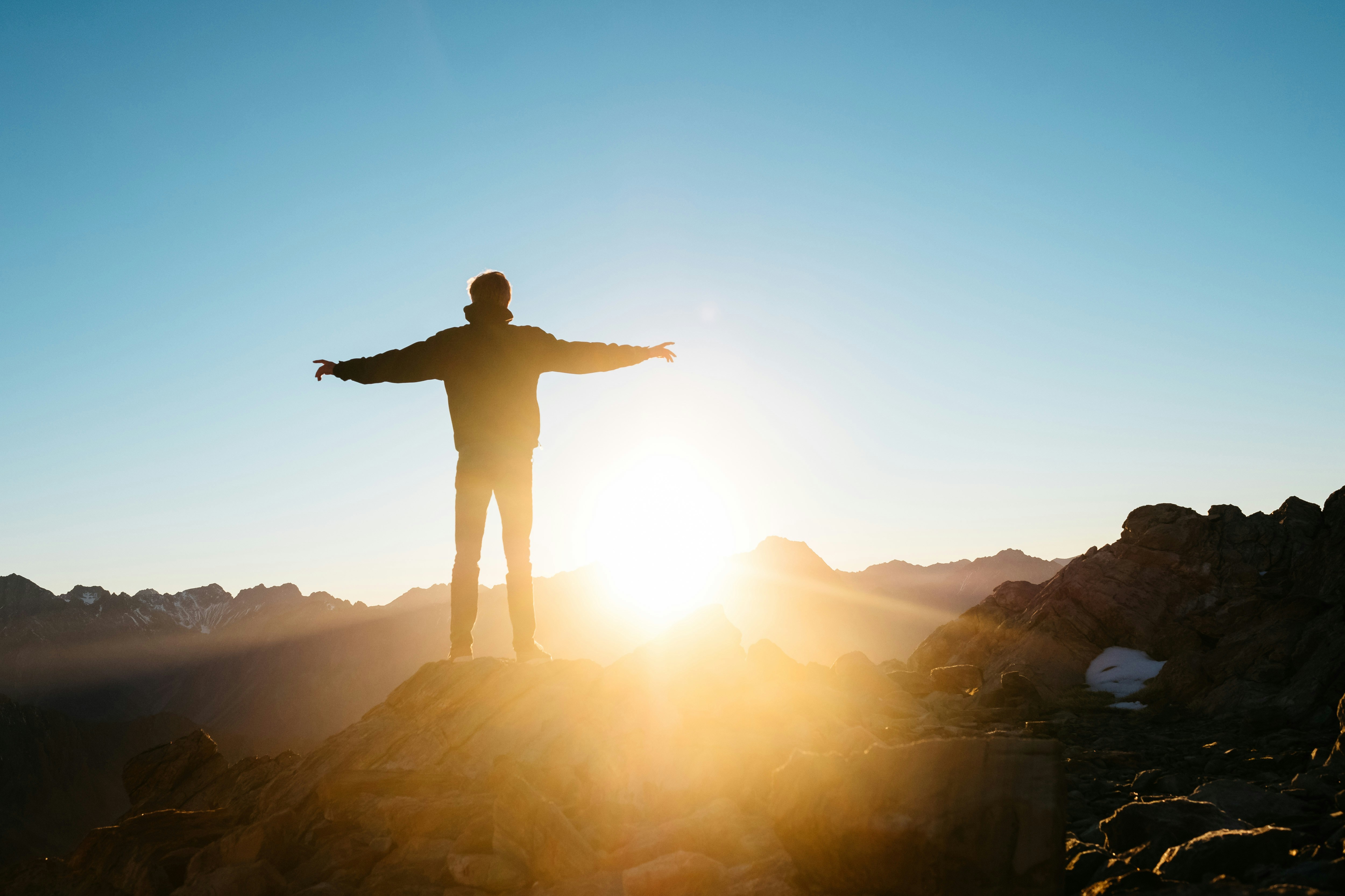 Silhouette of a person standing on a mountain peak with arms outstretched, facing a bright sunrise over a range of distant mountains and a clear sky.