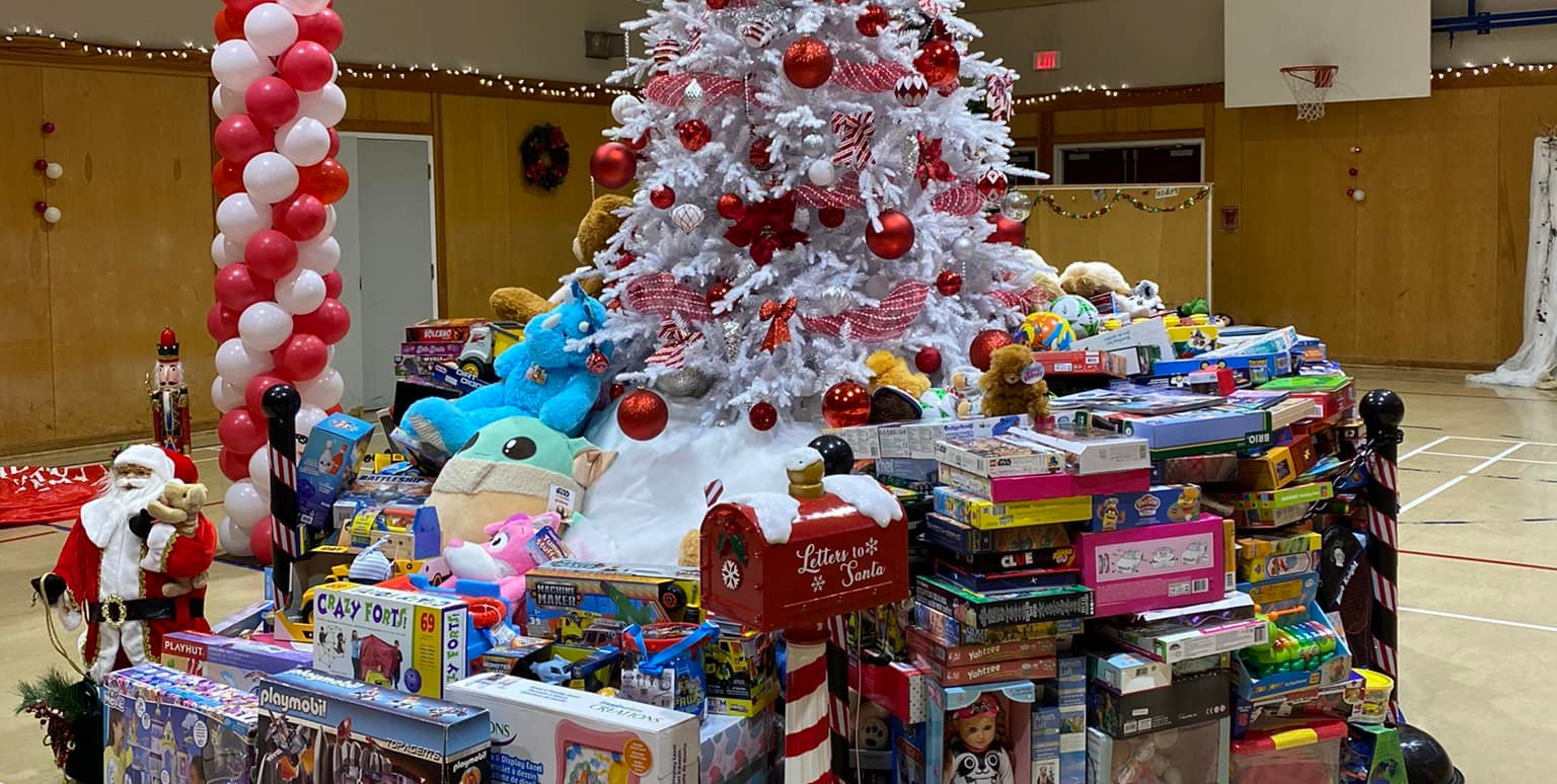 A large Christmas tree decorated with red and white ornaments stands indoors, surrounded by tables filled with toys, games, and stuffed animals, with festive decorations including balloons and holiday displays in the background.