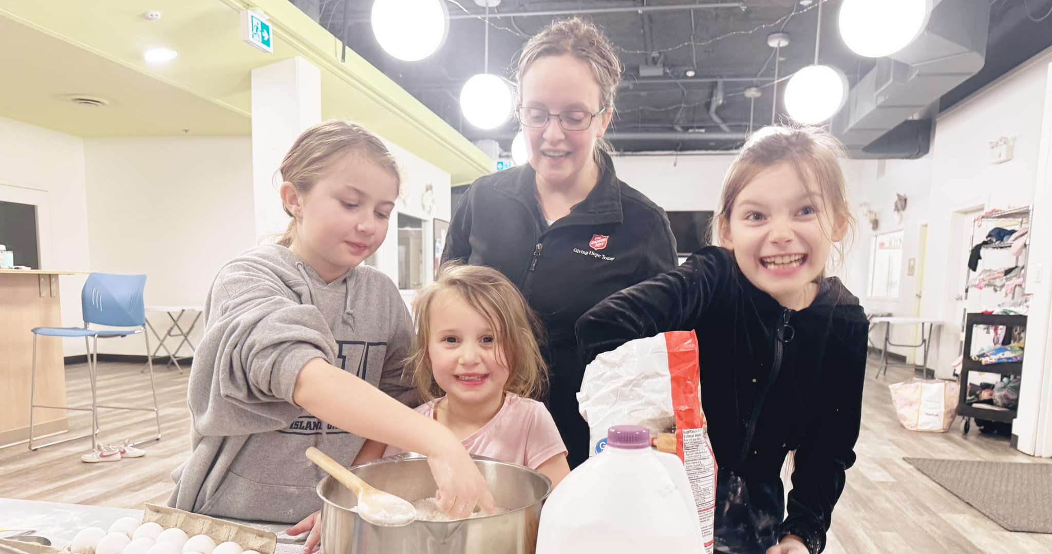 Children and an adult gathered around a kitchen counter mixing ingredients in a large bowl, with baking supplies such as flour, a carton, and utensils visible in a bright indoor space.
