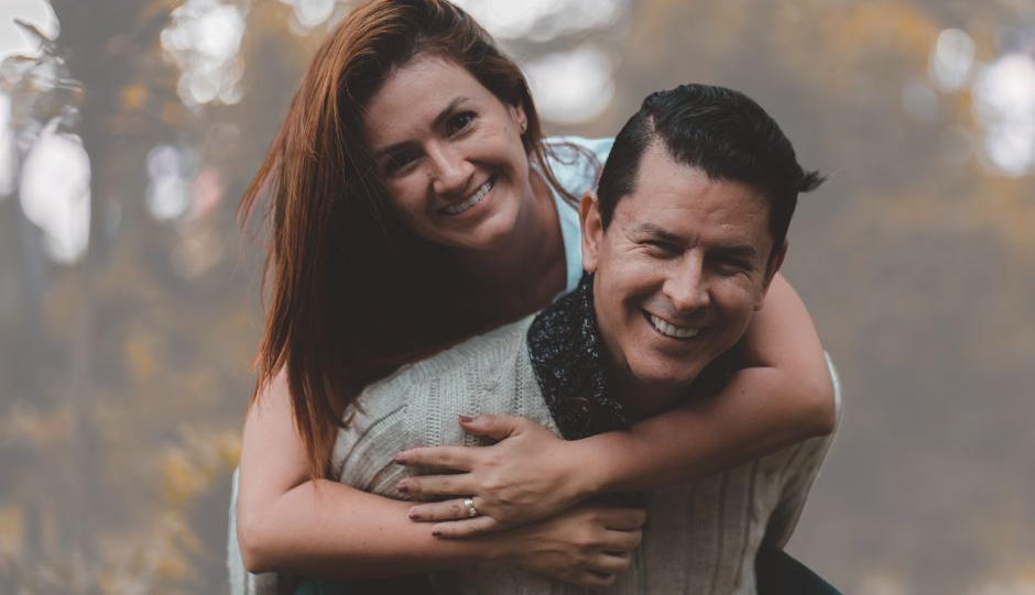A couple outdoors in a natural setting, with one person giving the other a piggyback ride against a backdrop of trees and soft natural light.