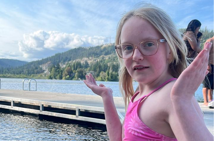 Child standing at the edge of a dock by a lake with arms raised, with water, a swimming ladder, and forested hills in the background during a beach event for the Partners in Mission plunge fundraiser.