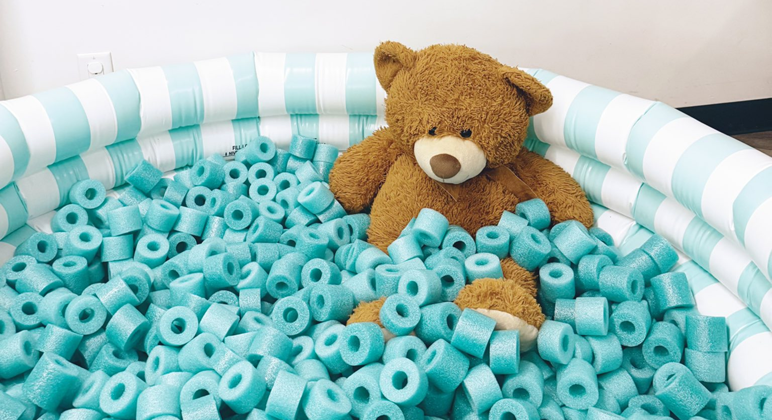 A small inflatable sensory play pool filled with turquoise foam pieces, with a brown teddy bear sitting inside.